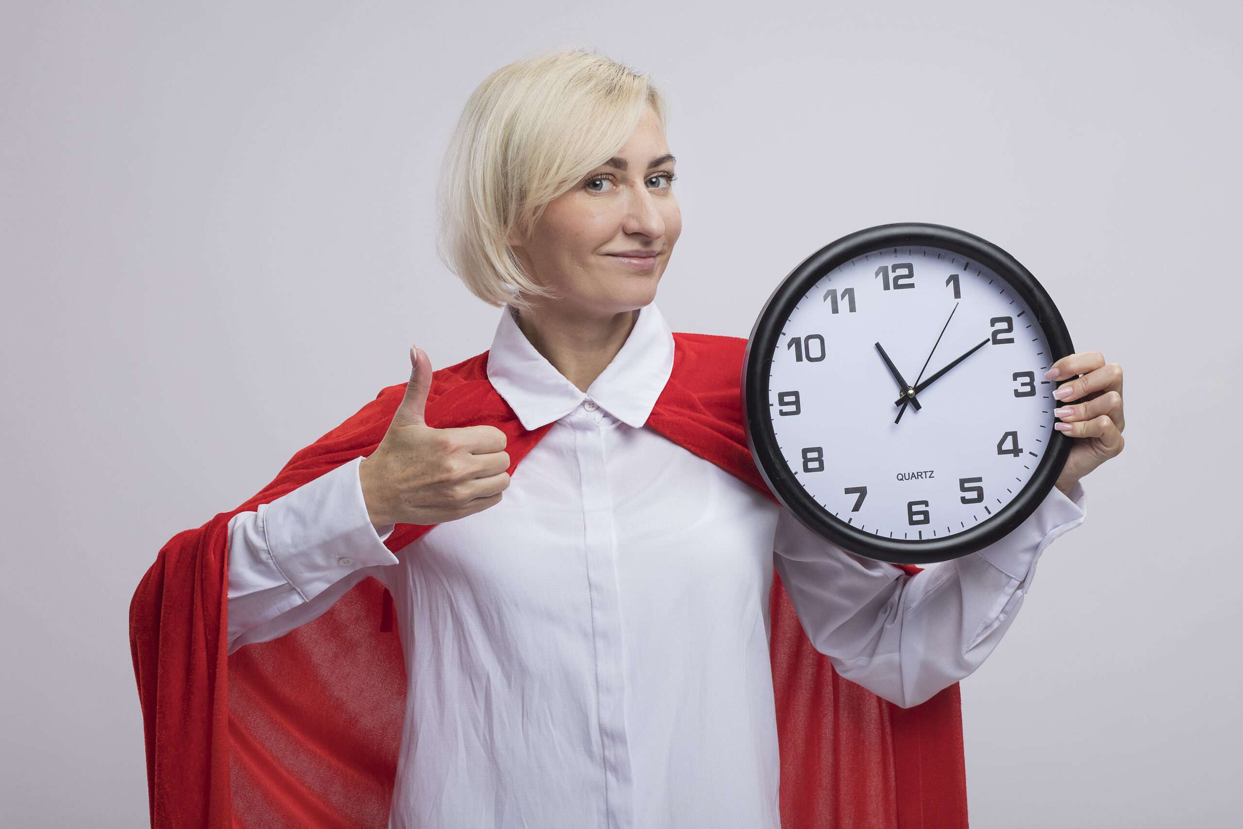 pleased middle-aged blonde superhero woman in red cape holding clock looking at camera showing thumb up isolated on white background